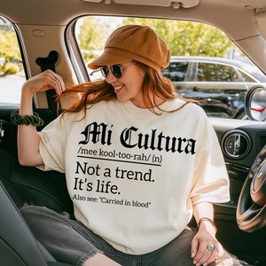 Cream sweatshirt with bold black text reading "Mi Cultura /mee kool-too-rah/ (n) Not a trend. It’s life. Also see: 'Carried in blood'" in Gothic and serif fonts, worn by a smiling woman in a car wearing sunglasses and a brown cap.