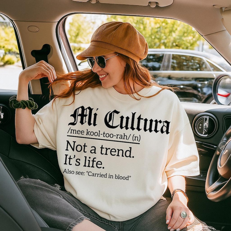 Cream sweatshirt with bold black text reading "Mi Cultura /mee kool-too-rah/ (n) Not a trend. It’s life. Also see: 'Carried in blood'" in Gothic and serif fonts, worn by a smiling woman in a car wearing sunglasses and a brown cap.