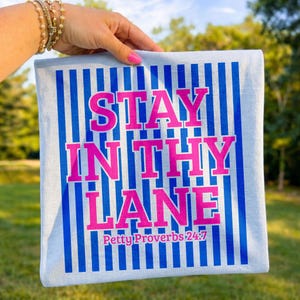 May include: A light blue square towel with vertical blue stripes. The towel features the text "STAY IN THY LANE" in bright pink, with "Petty Proverbs 24:7" below. The towel is held up by a person.