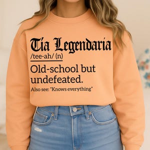 Light orange sweatshirt with bold black text reading "Tia Legendaria /tee-ah/ (n) Old-school but undefeated. Also see: 'Knows everything'" in Gothic and serif fonts.