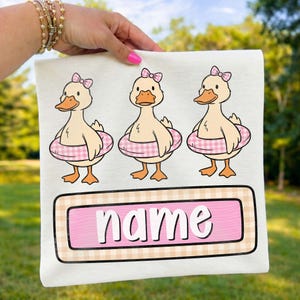 Puede incluir: Camiseta blanca con tres patos de dibujos animados con flotadores y lazos rosas. Los patos tienen picos y patas naranjas. Debajo, un rectángulo a cuadros rosa y beige con la palabra "name".