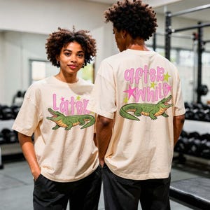 Indoor gym mockup showing two models wearing neutral beige oversized t-shirts with a colorful alligator graphic design. The front reads “Later” while the back displays “After While” in bold pink lettering with star accents. The modern fitness environment contrasts with the playful design, highlighting versatility for everyday casual wear and streetwear styling.