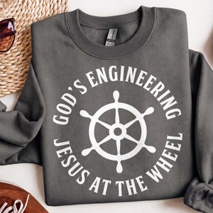 May include: A dark grey crewneck sweatshirt featuring a white graphic print of a ship's wheel centred within a circular text design. The text reads "GOD'S ENGINEERING" arched above the wheel and "JESUS AT THE WHEEL" arched below the wheel.