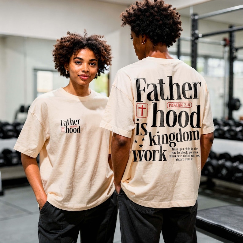 A man and a woman with curly afro hairstyles pose together in a gym setting. The woman stands facing forward, showing a small front chest print that reads "Father hood" on a cream t-shirt. The man stands facing away to display the large back print reading "Father hood is kingdom work" in bold black letters with red accents. Both wear black pants, showcasing a versatile, unisex streetwear look for couples.