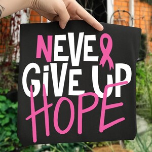 May include: Black tote bag with the words "NEVER GIVE UP HOPE" in white and pink lettering. A pink ribbon graphic is also featured. The bag is held up by a hand.