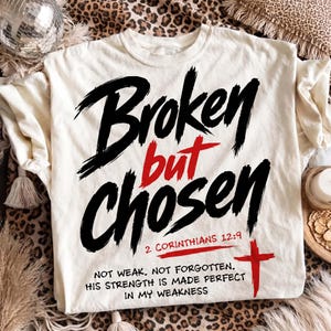 Cream T-shirt laid flat on a leopard print rug with a disco ball, tassel throw, and cozy neutral decor around it. The shirt features a bold Christian streetwear graphic reading “Broken but Chosen” in large black brush lettering, with “but,” the scripture reference, and cross accent in red. The lower quote about strength made perfect in weakness adds a powerful faith-based message.