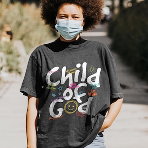 Lifestyle mockup of a woman walking outdoors wearing a dark charcoal “Child of God” Christian graphic t-shirt. The design includes multicolor typography with faith-based phrases, hearts, stars, and a smiley face detail, styled casually with jeans, showcasing modern inspirational Christian apparel in a natural city setting.