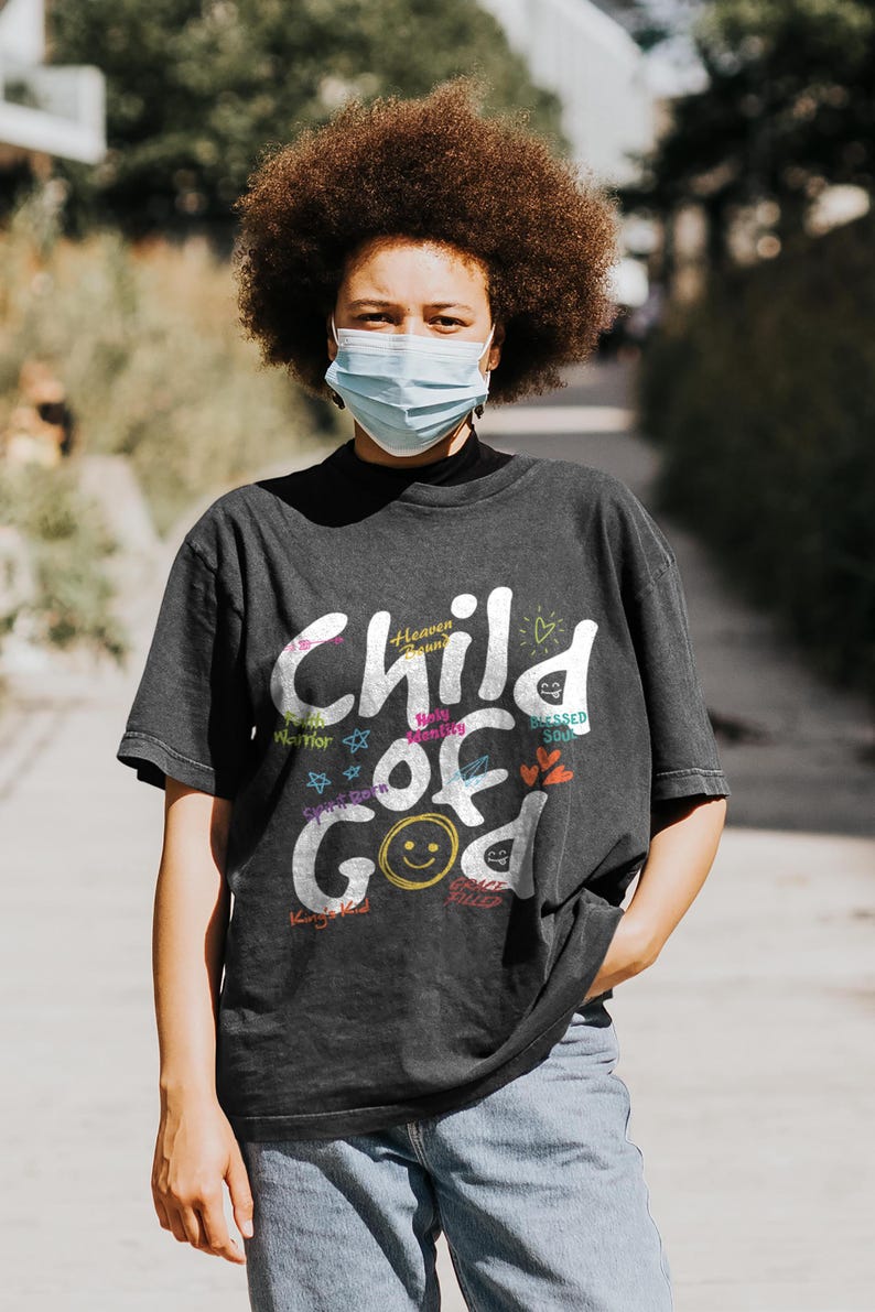 Lifestyle mockup of a woman walking outdoors wearing a dark charcoal “Child of God” Christian graphic t-shirt. The design includes multicolor typography with faith-based phrases, hearts, stars, and a smiley face detail, styled casually with jeans, showcasing modern inspirational Christian apparel in a natural city setting.