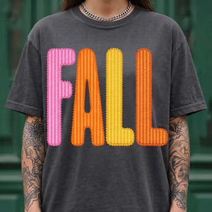 Op de afbeelding: Een antracietgrijs T-shirt met korte mouwen en het woord FALL in grote, verticaal gestreepte letters. De letter F is roze, de A is oranje, de L is geel en de tweede L is oranje. Elke letter heeft een geschulpte, gestikte rand.