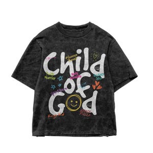 Dark washed black t-shirt shown in a flat lay product mockup against a white background, displaying large white “Child of God” text with playful Christian-themed words, hearts, stars, and graphic accents. The isolated product image clearly presents the vibrant faith-based shirt design for online listings.