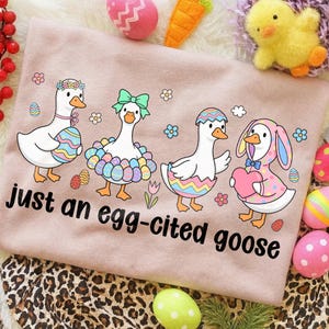 May include: A light pink t-shirt with the phrase "just an egg-cited goose" in black. The shirt features four cartoon geese decorated with Easter egg designs, flowers, and bows. Easter eggs and other decorations surround the shirt.