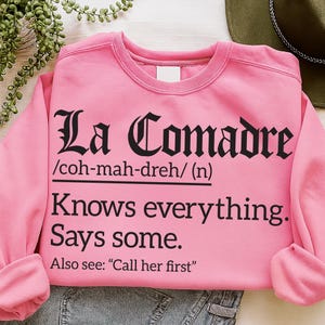 Pink sweatshirt with black text reading “La Comadre /coh-mah-dreh/ (n) Knows everything. Says some. Also see: ‘Call her first,’” laid flat with denim jeans and a hat nearby.