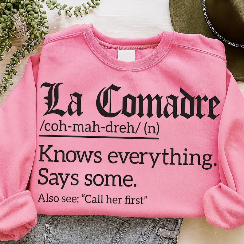 Pink sweatshirt with black text reading “La Comadre /coh-mah-dreh/ (n) Knows everything. Says some. Also see: ‘Call her first,’” laid flat with denim jeans and a hat nearby.