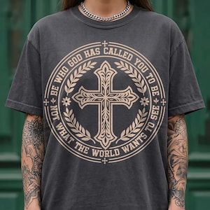 Close-up of a model with tattooed arms wearing a vintage washed charcoal gray graphic shirt and silver chain. The front displays a beige circular crest with a detailed cross, laurel leaves, and the quote "Be who God has called you to be not what the world wants to see". An edgy, stylish faith-based graphic tee option for modern streetwear fashion.