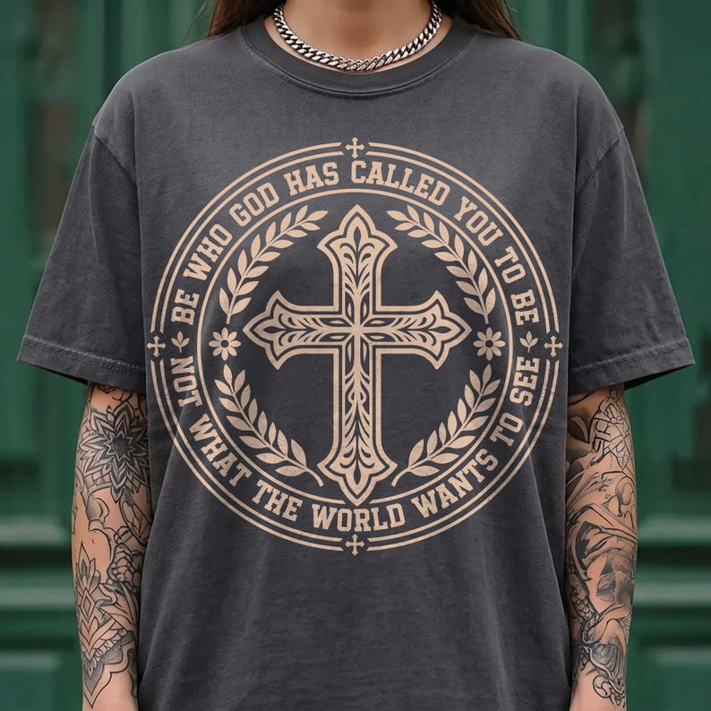 Close-up of a model with tattooed arms wearing a vintage washed charcoal gray graphic shirt and silver chain. The front displays a beige circular crest with a detailed cross, laurel leaves, and the quote "Be who God has called you to be not what the world wants to see". An edgy, stylish faith-based graphic tee option for modern streetwear fashion.