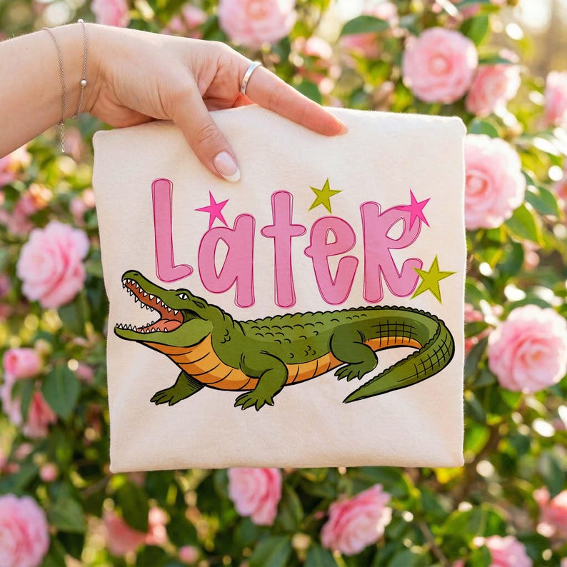 A neatly folded cream t-shirt displayed in a hand against a soft floral backdrop, featuring the word “Later” in pink bubble letters with bright star accents and a detailed green alligator graphic. The composition highlights the clean print quality and trendy aesthetic, perfect for showcasing digital design mockups for summer apparel or casual wear collections.