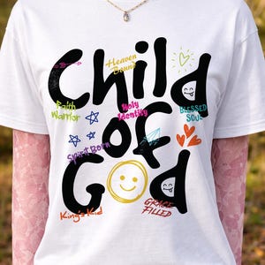 White t-shirt on a mannequin styled with pink lace sleeves and a delicate necklace, showcasing a bold black “Child of God” Christian graphic with colorful handwritten faith phrases and doodle icons. This outdoor mockup emphasizes feminine styling and cheerful inspirational church apparel.