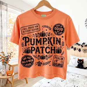 May include: An orange short-sleeve t-shirt featuring black graphic text and pumpkin illustrations. The design includes the words PUMPKIN PATCH, I WILL PRAISE HIM IN EVERY SEASON, GOD IS GOOD ALL THE TIME, ARE YOU FALL-O-WEEN JESUS, and PSALM 23.