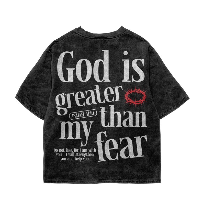 Flat lay presentation of a vintage washed charcoal gray t-shirt spread flat on a clean white background. The shirt features prominent white typography reading "God is greater than my fear" paired with a red crown of thorns accent and the verse "Isaiah 41:10". A minimalist, powerful faith graphic tee providing comfort, hope, and inspirational scripture fashion.