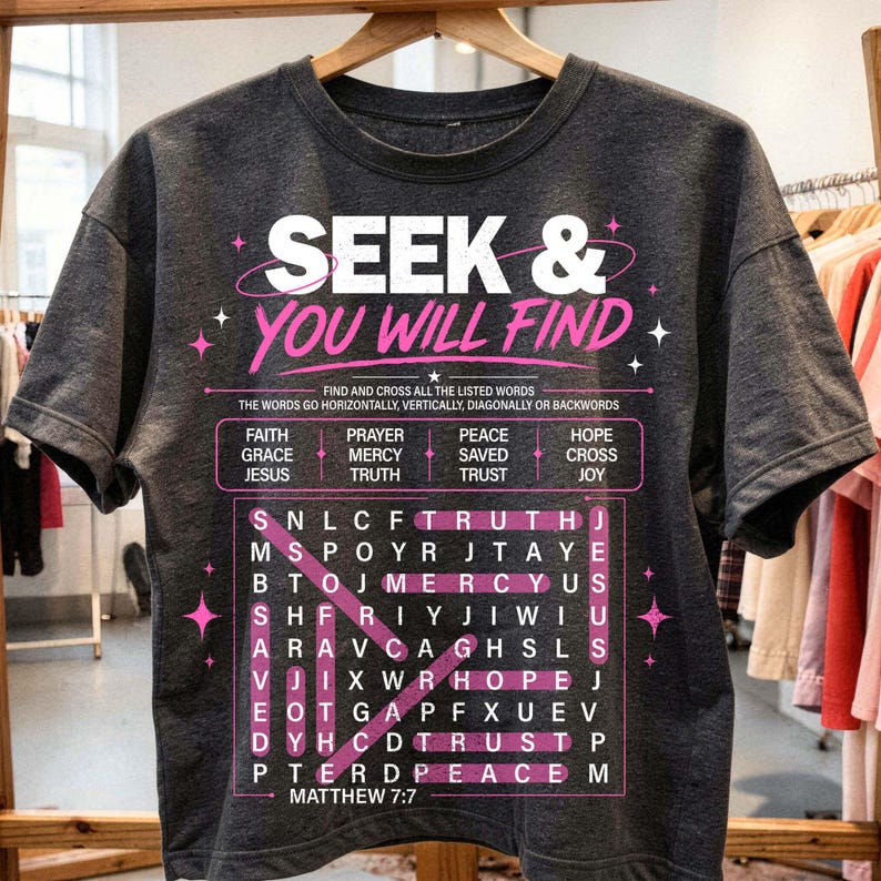 Dark charcoal T-shirt hanging on a wooden hanger in a boutique clothing shop, featuring a bold Christian “Seek & You Will Find” design in white and pink. The artwork includes a faith word search puzzle, highlighted scripture words, Matthew 7:7 reference, and star accents, giving the shirt a trendy streetwear look for faith-based apparel. Ideal mockup for Etsy listings.