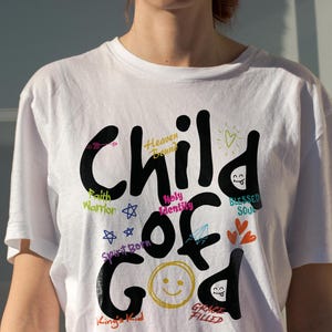 White Christian t-shirt worn in natural sunlight, featuring bold black “Child of God” lettering with bright accent words like Heaven Bound, Holy Identity, Faith Warrior, Blessed Soul, and Grace Filled. The clean lifestyle mockup highlights uplifting faith typography and colorful doodle elements for church apparel.