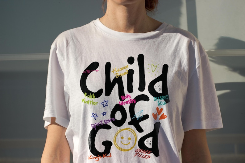 White Christian t-shirt worn in natural sunlight, featuring bold black “Child of God” lettering with bright accent words like Heaven Bound, Holy Identity, Faith Warrior, Blessed Soul, and Grace Filled. The clean lifestyle mockup highlights uplifting faith typography and colorful doodle elements for church apparel.