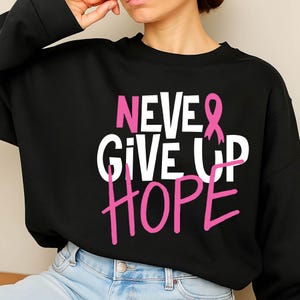 May include: Black sweatshirt with the words "NEVER GIVE UP HOPE" in white and pink lettering. A pink ribbon graphic is incorporated into the design. The sweatshirt is a crew neck style.