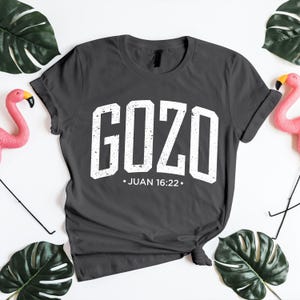 Flat lay mockup of a dark charcoal gray unisex t-shirt featuring the bold distressed word “GOZO” in white varsity-style lettering, with the Bible reference Juan 16:22 centered below. The shirt is styled with a casual knotted hem on a bright white background, accented by green tropical leaves and pink flamingo decor. This inspirational Spanish Christian t-shirt mockup highlights faith-based typography, modern apparel styling, and a clean presentation ideal for showcasing religious designs.