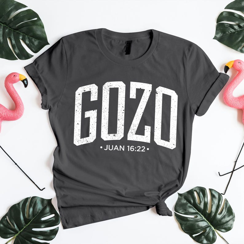 Flat lay mockup of a dark charcoal gray unisex t-shirt featuring the bold distressed word “GOZO” in white varsity-style lettering, with the Bible reference Juan 16:22 centered below. The shirt is styled with a casual knotted hem on a bright white background, accented by green tropical leaves and pink flamingo decor. This inspirational Spanish Christian t-shirt mockup highlights faith-based typography, modern apparel styling, and a clean presentation ideal for showcasing religious designs.