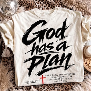 Cream T-shirt laid flat on a leopard print rug with a disco ball, tasseled blanket, and cozy neutral decor around it. The shirt features a bold Christian graphic reading “God has a Plan” in large black brush lettering, accented with a red cross, Jeremiah 29:11 reference, and a shortened scripture quote near the hem for an edgy faith-based streetwear style.
