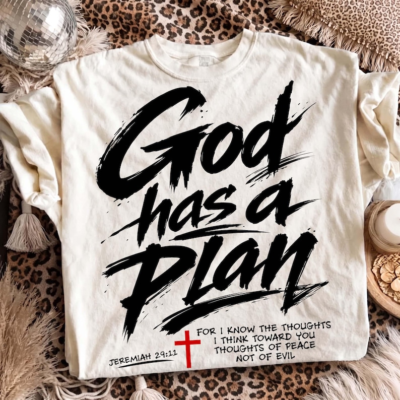 Cream T-shirt laid flat on a leopard print rug with a disco ball, tasseled blanket, and cozy neutral decor around it. The shirt features a bold Christian graphic reading “God has a Plan” in large black brush lettering, accented with a red cross, Jeremiah 29:11 reference, and a shortened scripture quote near the hem for an edgy faith-based streetwear style.