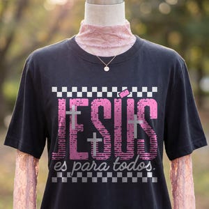 Black women’s t-shirt displayed on a mannequin layered over pink lace sleeves with a delicate gold necklace, featuring vibrant pink “JESÚS” lettering, subtle cross overlays, and checkerboard accents framing “es para todos.” This stylish Spanish Christian graphic tee merges modern streetwear elements with meaningful faith expression.