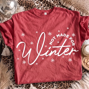 May include: A red short-sleeved t-shirt featuring white text that reads "NOT MADE FOR Winter" in a mix of block and script fonts. The design is accented with various white snowflake illustrations scattered across the front of the garment.