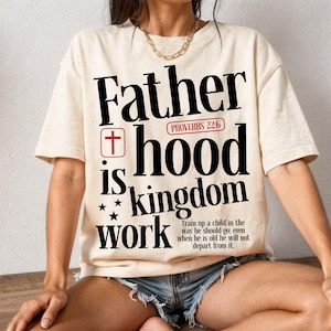 A woman sits cross-legged on a wooden floor against a white textured wall, wearing an oversized cream t-shirt, distressed denim shorts, and a gold chain necklace. The front of the shirt features a large Christian typographic print reading "Father hood is kingdom work" in stylized black letters. The design includes a red cross symbol, a red Proverbs 22:6 tag, stars, and the small scripture text at the bottom.