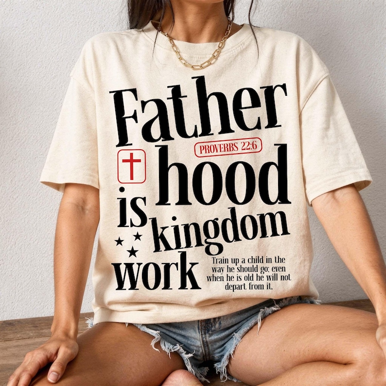 A woman sits cross-legged on a wooden floor against a white textured wall, wearing an oversized cream t-shirt, distressed denim shorts, and a gold chain necklace. The front of the shirt features a large Christian typographic print reading "Father hood is kingdom work" in stylized black letters. The design includes a red cross symbol, a red Proverbs 22:6 tag, stars, and the small scripture text at the bottom.