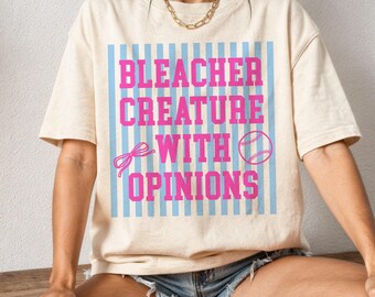Op de afbeelding: Crèmekleurig T-shirt met de roze tekst "Bleacher Creature With Opinions". Het ontwerp bevat een honkbal, een strik en blauwe verticale strepen. Het shirt heeft korte mouwen en een losse pasvorm.