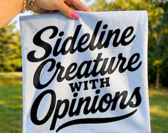 Op de afbeelding: Een lichtblauwe vierkante handdoek met de tekst "Sideline Creature With Opinions" in zwarte letters. De handdoek wordt vastgehouden door een hand met roze nagellak, met een vage groene achtergrond.
