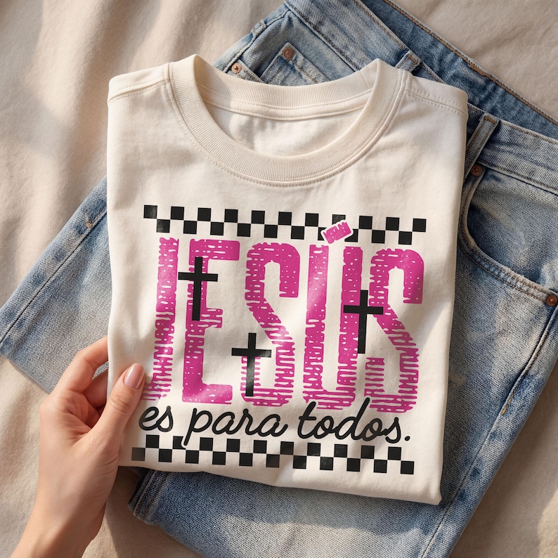 Cream Christian t-shirt folded over light wash denim jeans featuring bold pink distressed “JESÚS” lettering with integrated black crosses, checkerboard stripe accents, and the phrase “es para todos.” in script below. Styled in soft natural lighting with a hand holding the shirt, this faith-based Spanish graphic tee blends retro streetwear aesthetics with inspirational church apparel.