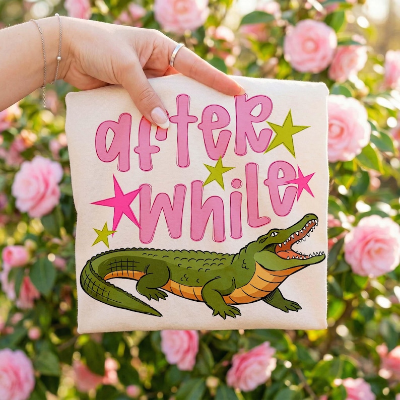 Close-up of a folded cream-colored t-shirt held by a hand in a floral garden setting, showcasing a vibrant “After While” design with bold pink lettering, colorful stars, and a detailed cartoon alligator illustration. The soft natural lighting and blooming background enhance the playful and stylish vibe, making it ideal for boutique fashion and trendy graphic tee lovers.