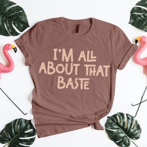 Brown Thanksgiving T-shirt with “I’m All About That Baste” in beige vintage font. Funny holiday cooking shirt for chefs, moms, or food lovers celebrating turkey season. Perfect Thanksgiving outfit for family dinner photos or Friendsgiving parties. Playful fall apparel and Thanksgiving PNG graphic for crafters or sublimation printing projects.
