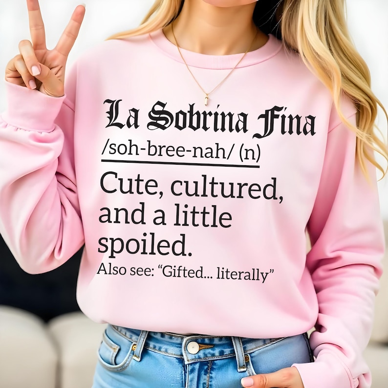 Light pink sweatshirt with black text reading “La Sobrina Fina /soh-bree-nah/ (n) Cute, cultured, and a little spoiled. Also see: ‘Gifted… literally,’” modeled by a woman making a peace sign.