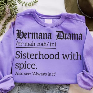 Lavender sweatshirt with black text reading “Hermana Drama /er-mah-nah/ (n) Sisterhood with spice. Also see: ‘Always in it,’” laid flat with casual styling beside a hat and greenery.