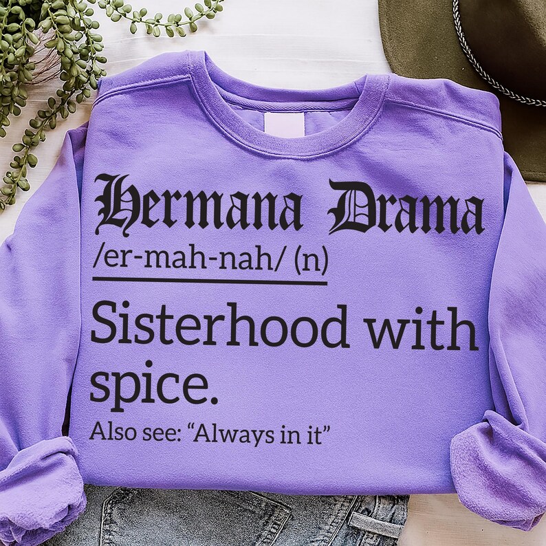 Lavender sweatshirt with black text reading “Hermana Drama /er-mah-nah/ (n) Sisterhood with spice. Also see: ‘Always in it,’” laid flat with casual styling beside a hat and greenery.