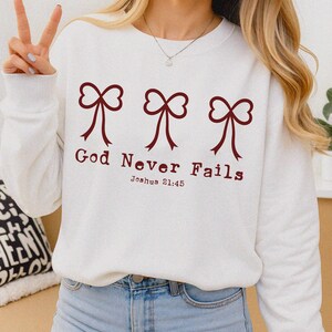 May include: White sweatshirt with three maroon bow graphics above the text "God Never Fails" and the Bible verse Joshua 21:45. The sweatshirt is a casual, comfortable style.