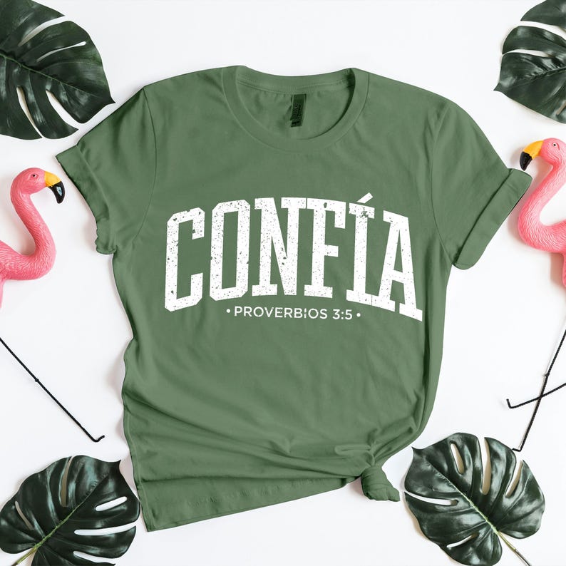 Minimal flat lay of a sage green unisex Christian t-shirt displaying the word “CONFÍA” in bold white distressed collegiate lettering, paired with Proverbios 3:5 beneath. The shirt is knotted at the bottom for a relaxed look and photographed on a white background with decorative tropical leaves and pink flamingos. This mockup is ideal for presenting Spanish faith apparel, Bible verse designs, and inspirational Christian clothing with a modern, lifestyle aesthetic.
