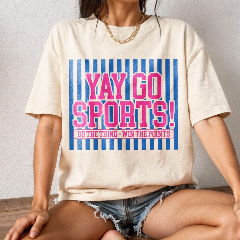 Lifestyle mockup of a woman seated wearing a relaxed fit cream t-shirt with distressed denim shorts, displaying a large pink and blue “Yay Go Sports!” varsity design with vertical stripe backdrop and “Do The Thing – Win The Points” text, perfect for showcasing a comfortable game day sports fan tee in natural lighting.