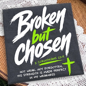 Dark folded apparel or canvas-style flat lay shown on a lace and woven rattan background. The design reads “Broken but Chosen” in oversized white brush lettering, with “but,” the cross, and 2 Corinthians 12:9 reference highlighted in bright green. The bottom message, “Not weak. Not forgotten. His strength is made perfect in my weakness,” gives the mockup a bold Christian streetwear feel.