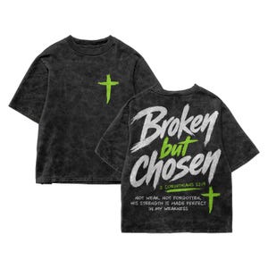 Black acid-wash oversized T-shirt mockup showing front and back views on a plain white background. The front has a small green cross chest print, while the back features a bold Christian streetwear design reading “Broken but Chosen” in large white brush lettering with green accents. The 2 Corinthians 12:9 reference, cross detail, and weakness-to-strength message create an edgy faith-based shirt mockup.