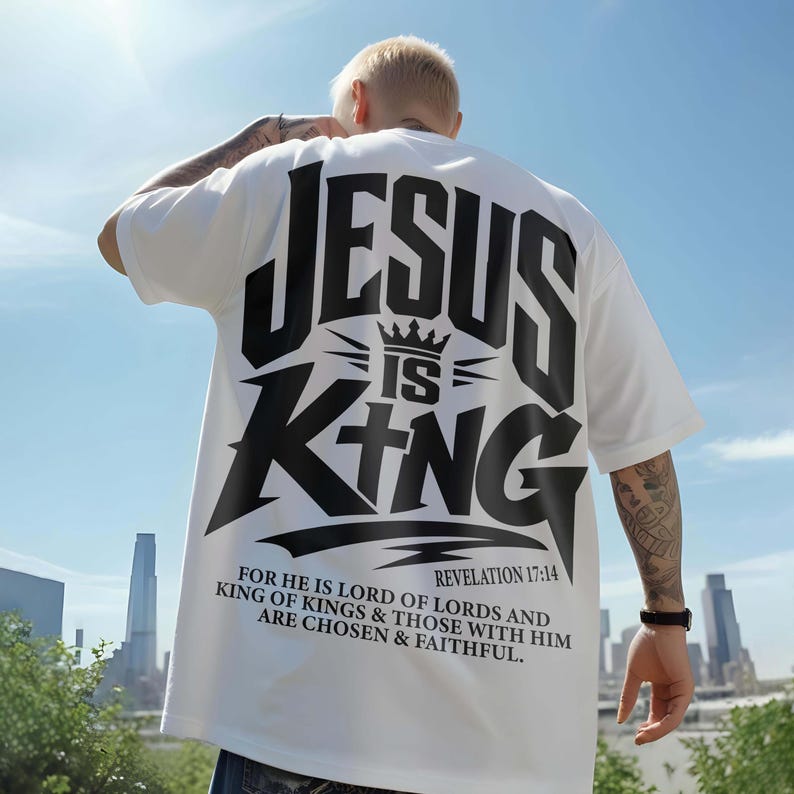Back view of a model in an oversized white graphic t-shirt against a clear sky and city background. The back features bold black typography stating "Jesus Is King" with a crown and cross motif, accompanied by "Revelation 17:14" and scripture. A modern Christian streetwear shirt designed for a bold urban aesthetic and daily faith expression.