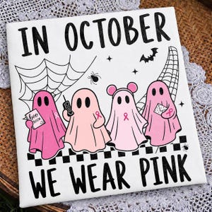 May include: A white square textile featuring the black text IN OCTOBER WE WEAR PINK. The design includes four pink ghosts, each with a pink ribbon symbol, positioned above a black and white checkered border, spiderwebs, a bat, and a spider.